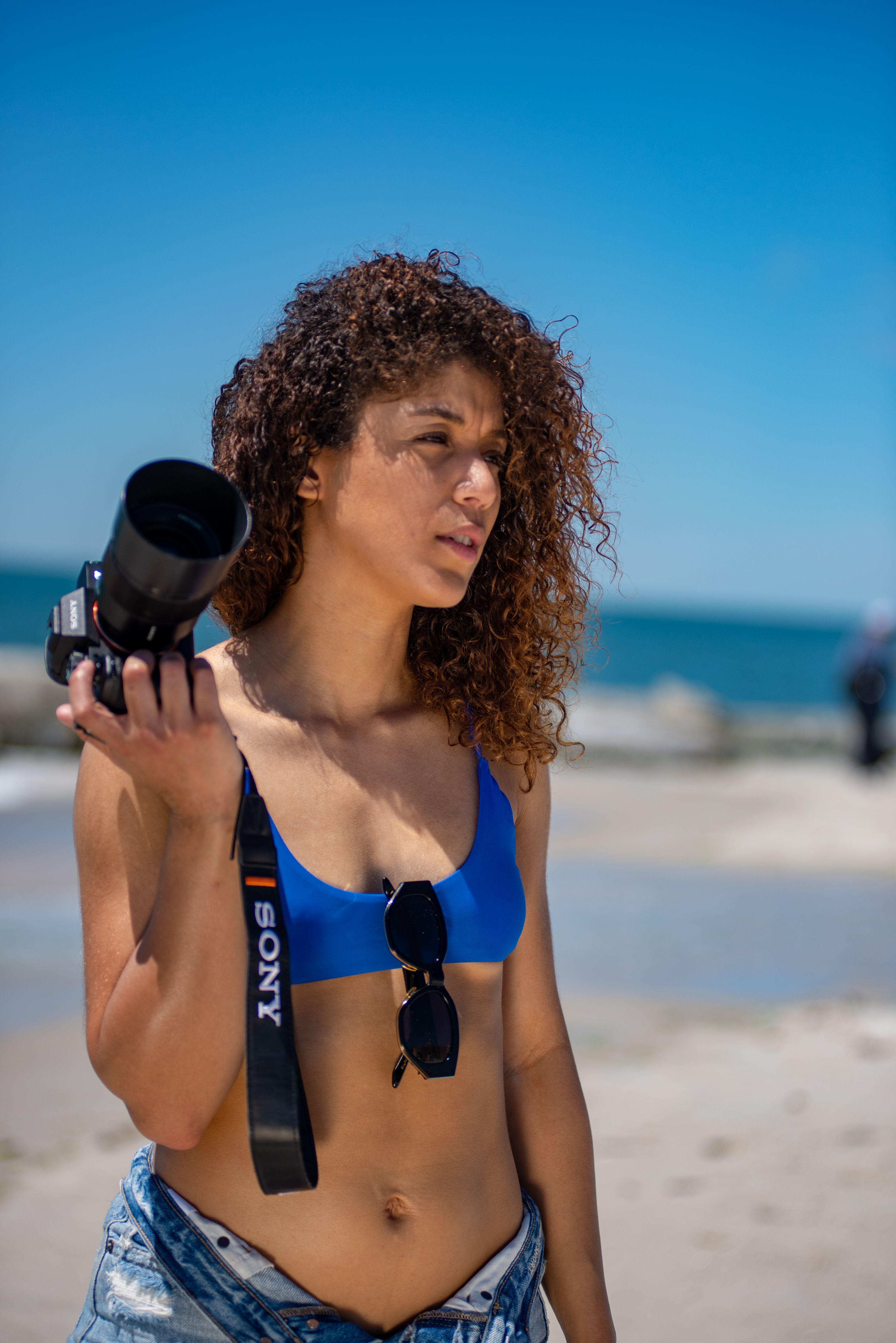 Stylish woman with curly hair holding a camera on Long Beach, NY, under a clear blue sky.