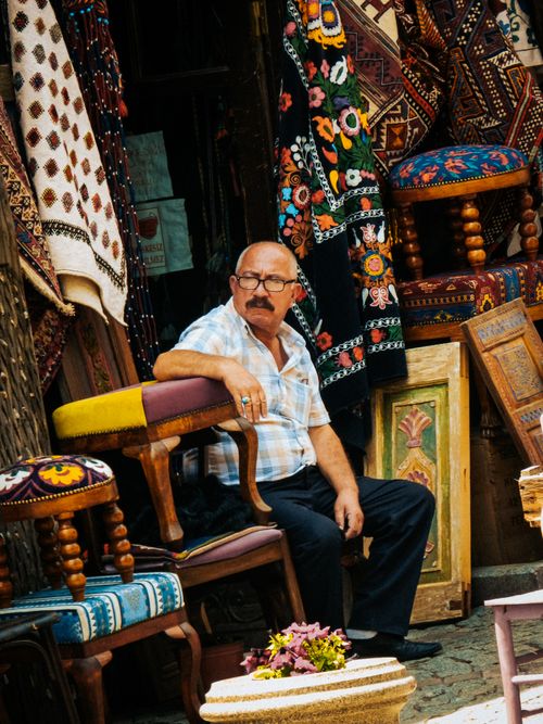 Free Senior vendor sitting by an array of colorful textiles and chairs in a market stall. Stock Photo