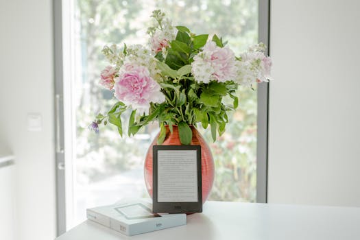 A vibrant floral arrangement in a red vase with a tablet on a table, indoors by a window.
