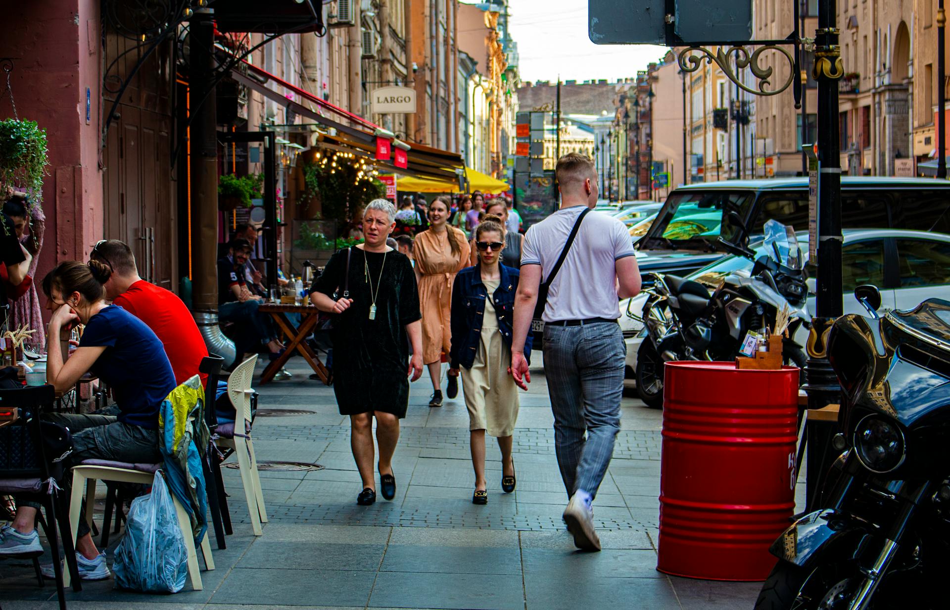 A lively street scene featuring people walking, outdoor cafés, and parked vehicles on a sunny day.