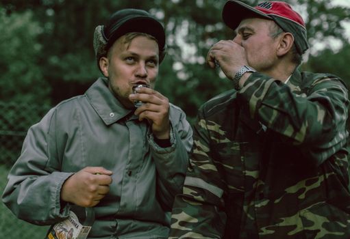 Two men in coats enjoying a drink in shot glasses outdoors, one in camouflage, sharing a moment.