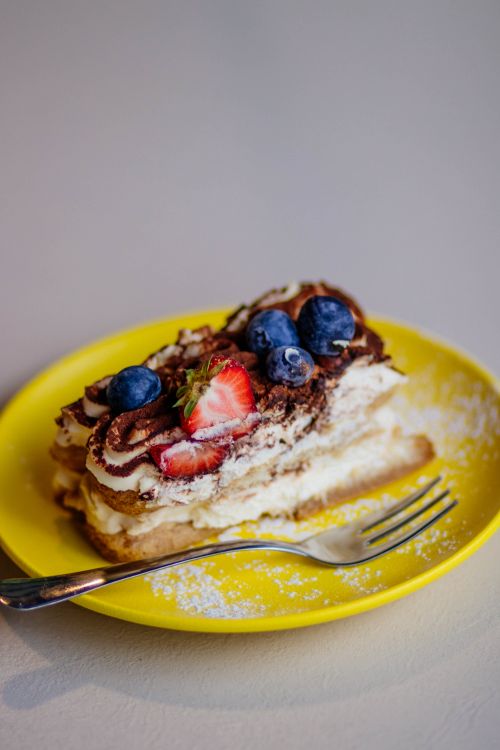 Free A slice of creamy cake topped with fresh strawberries and blueberries on a vibrant yellow plate. Stock Photo