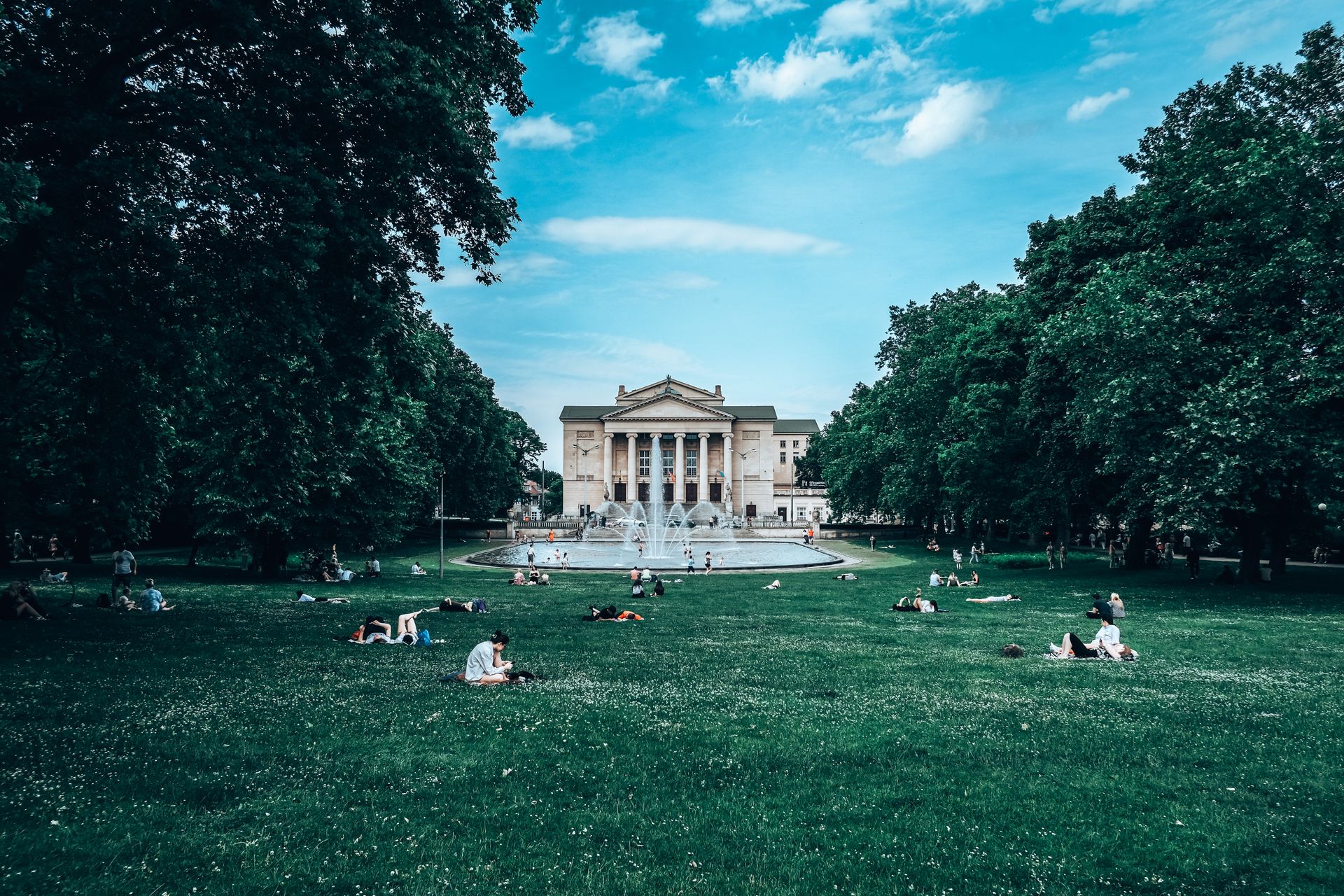 People enjoying a sunny day at Adam Mickiewicz Park in Poznan, Poland, with a historic building backdrop.