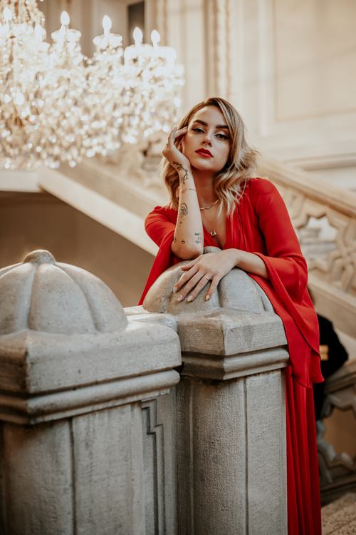 Free Tattooed woman in a red dress poses by a vintage staircase, exuding elegance and style. Stock Photo