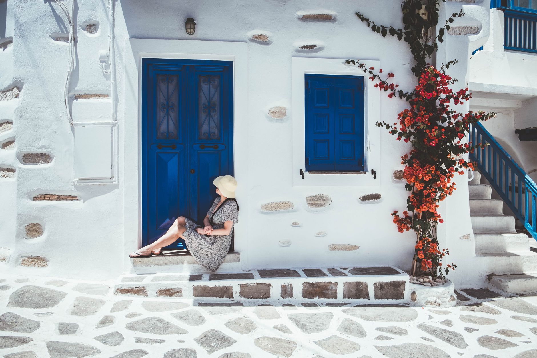 A woman in a sun hat rests by a blue door among flowers in Paros, Greece.