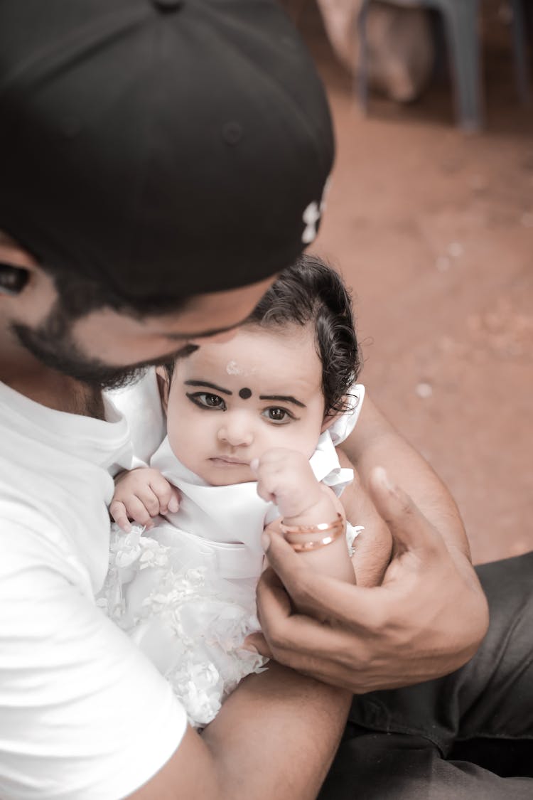 Photo Of A Baby In A Traditional Indian Makeup And White Clothing Being Held By A Man In His Arms