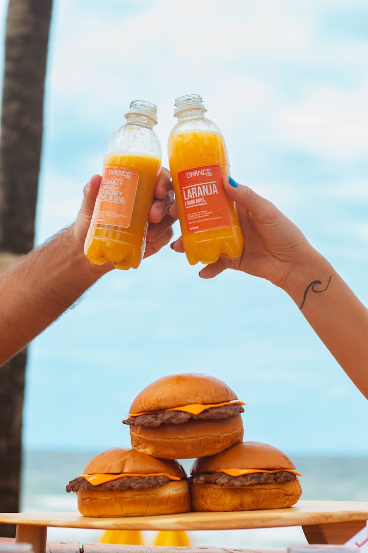 Close-up Of People Clinking Their Bottles With Juice Over Burgers 