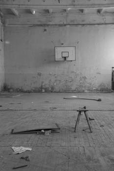 Black and white photo of a neglected indoor basketball court with peeling walls and scattered debris.