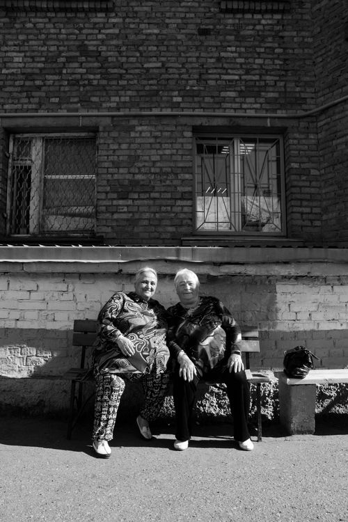 Free Two elderly women sitting on a bench against a brick wall outside, enjoying a sunny day. Stock Photo