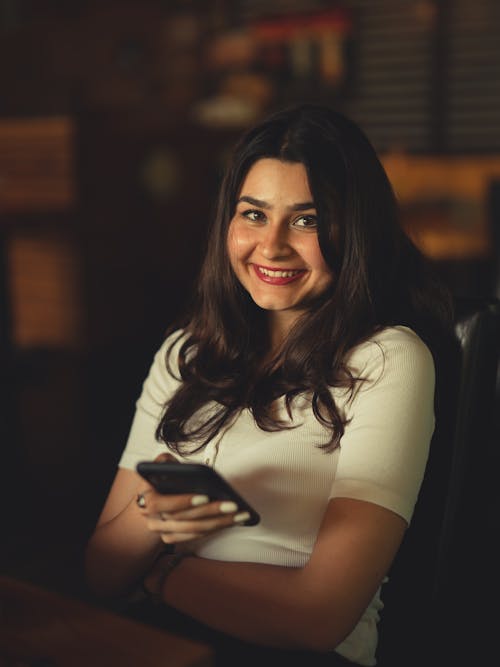 Free A Woman in White Shirt Holding a Cellphone while Smiling at the Camera Stock Photo