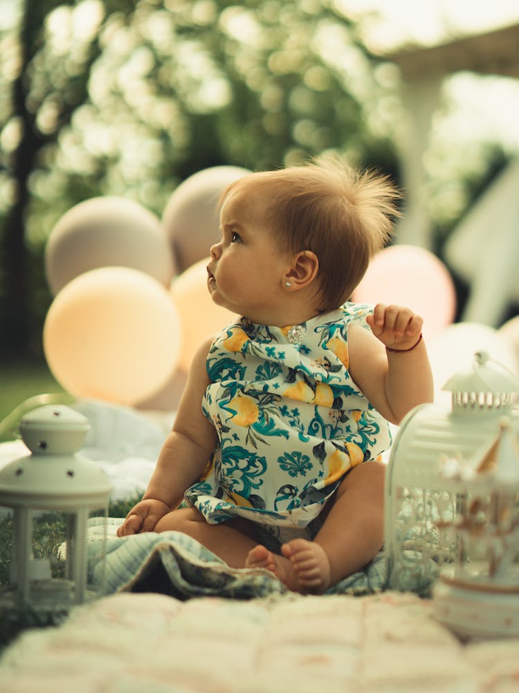 Baby Girl Wearing Patterned Clothing Sitting With Balloons In A Park