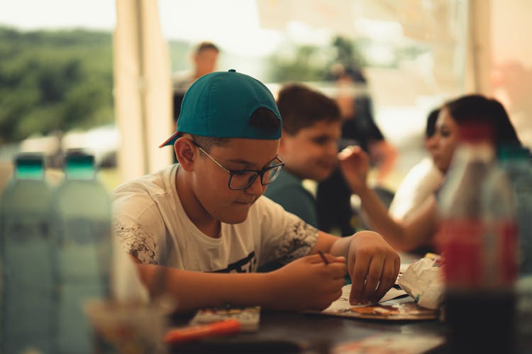 Boy In Hat And Glasses Sitting At Table In Cafe