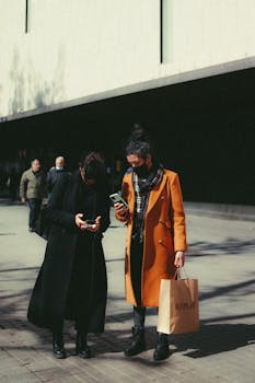 Two people in stylish winter coats using mobile phones on a city street.