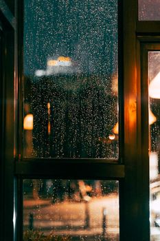 Rain-drenched window capturing urban night lights in a cozy ambiance.