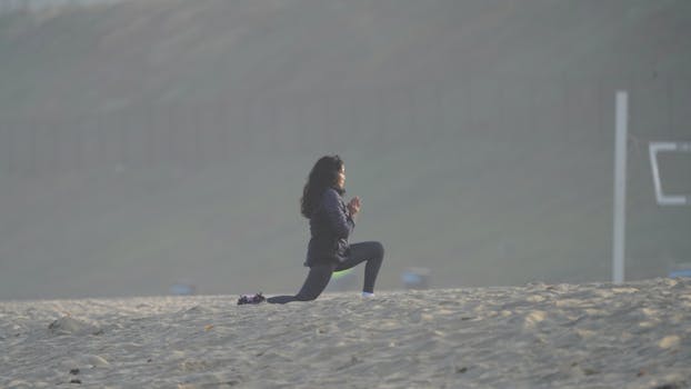 A woman kneels on a sandy beach, practicing yoga at dawn. Peaceful and serene atmosphere.