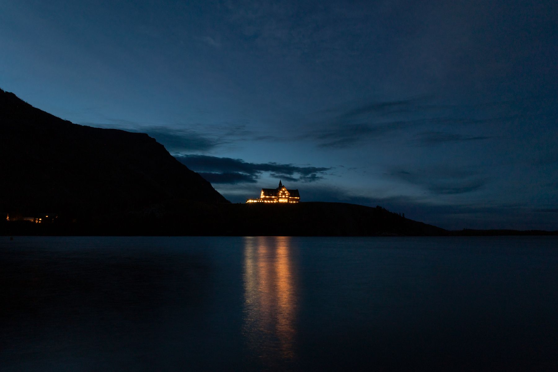 A still lake at blue hour, with one lit house on the far shore reflected in the water