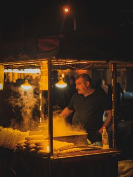 A man selling corncobs from a street food cart at night, illuminated by warm lighting.