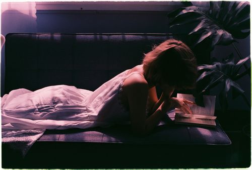 Free A woman lies on a couch in soft light, deeply engrossed in a book, creating a serene and introspective mood. Stock Photo