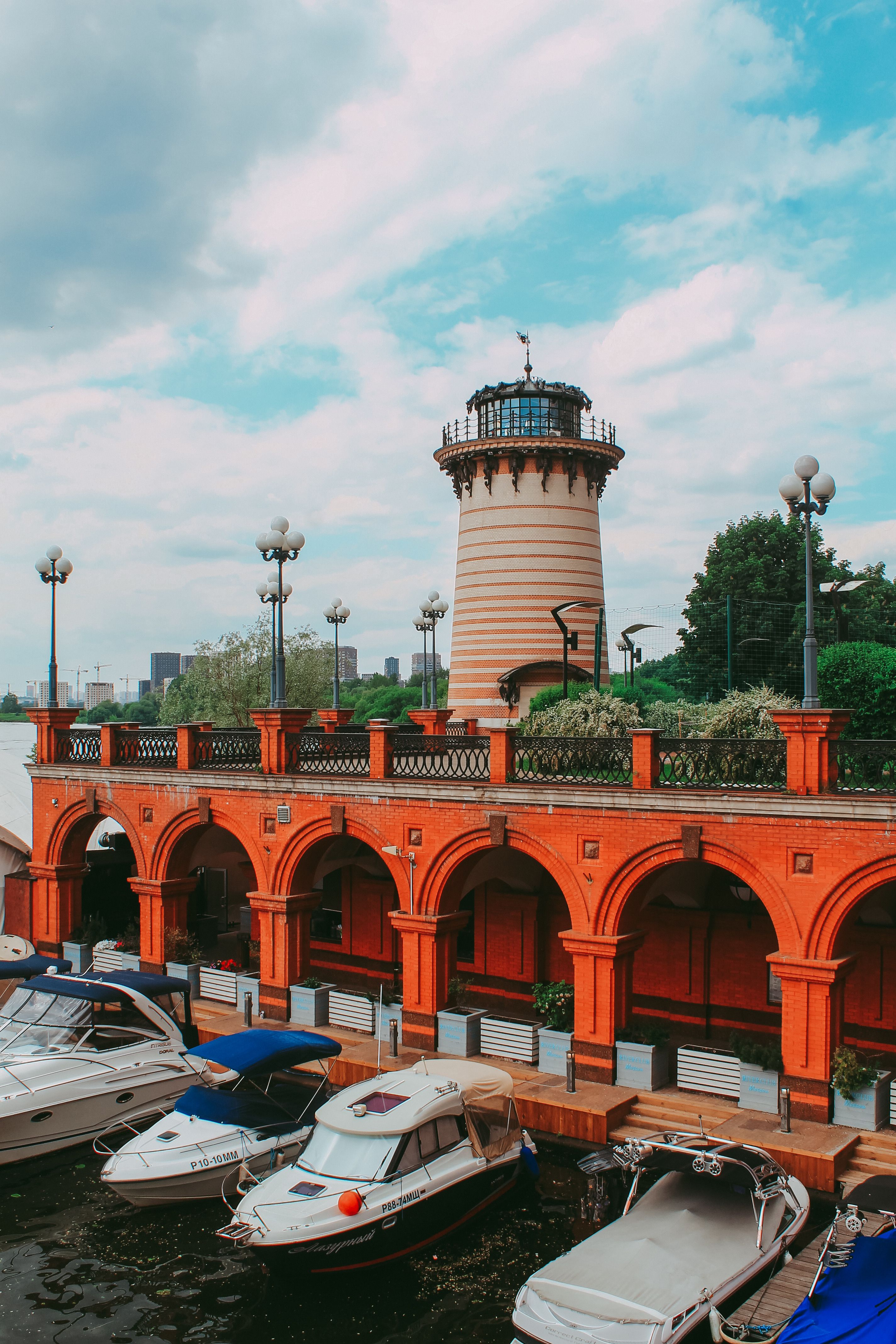 Scenic view of boats moored by a red brick pier with a lighthouse against a bright sky.