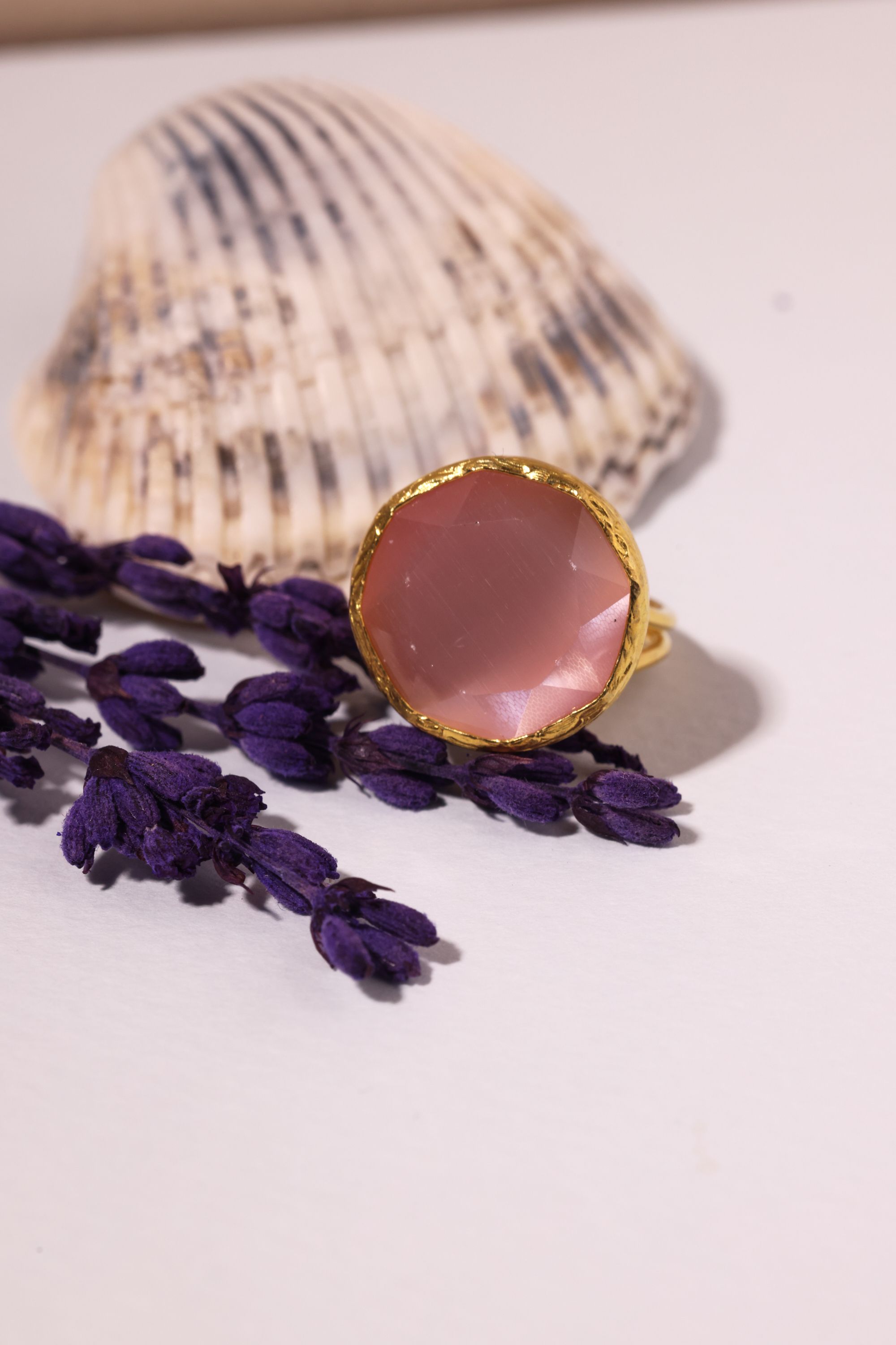 A close-up of a pink stone ring beside a shell and lavender on a white surface.