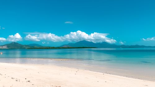 Free Peaceful beach scene with white sand, turquoise water, and mountain backdrop under a bright blue sky. Stock Photo