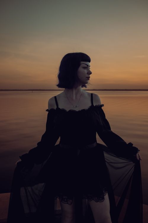 Free A woman in a black dress stands gracefully by the ocean at sunset, creating a serene silhouette. Stock Photo
