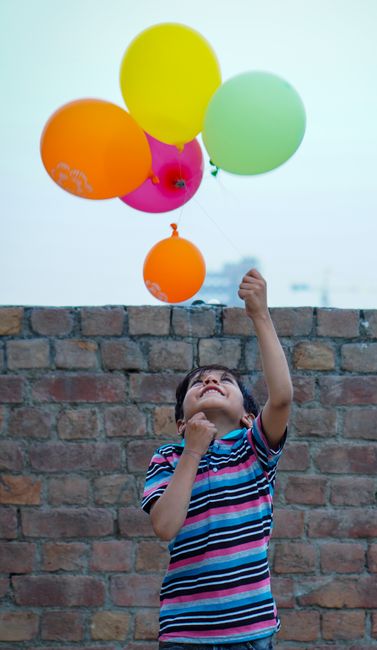 Young boy joyfully playing with colorful balloons against a brick wall in Pakistan.