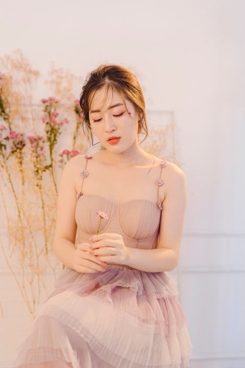 Free A graceful woman in a pastel dress holding a flower, exuding elegance and calmness indoors. Stock Photo