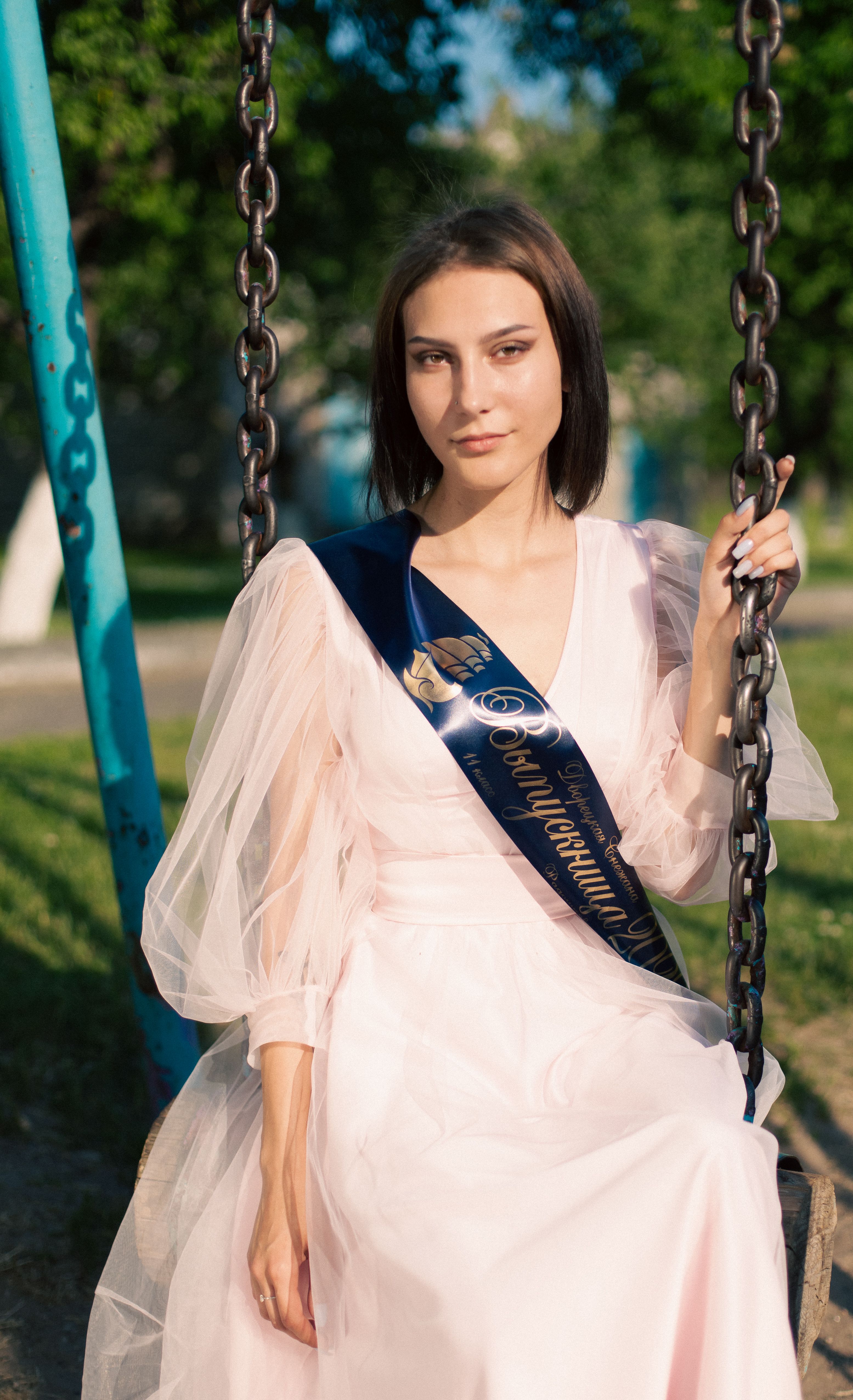 Young woman in a pink dress on a swing, posing gracefully outdoors on a sunny day.
