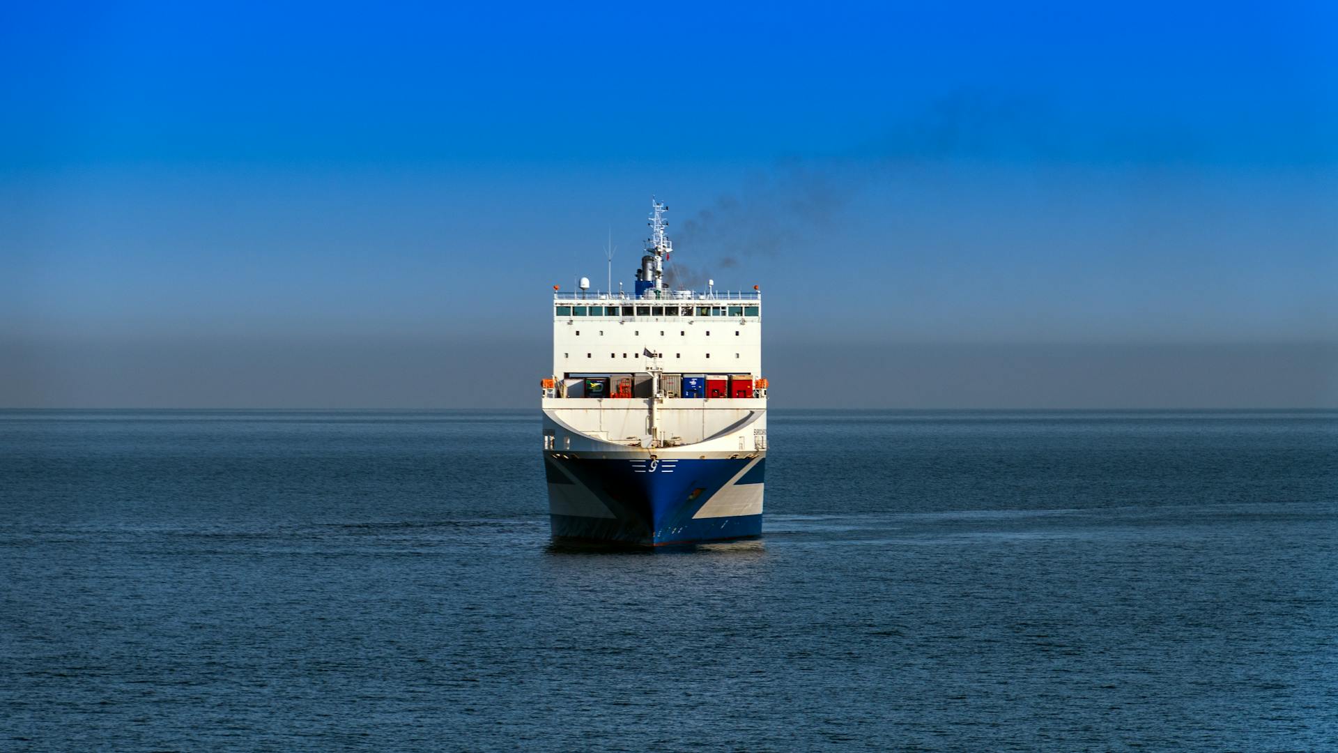 A large cargo vessel travels across the ocean under a clear blue sky.