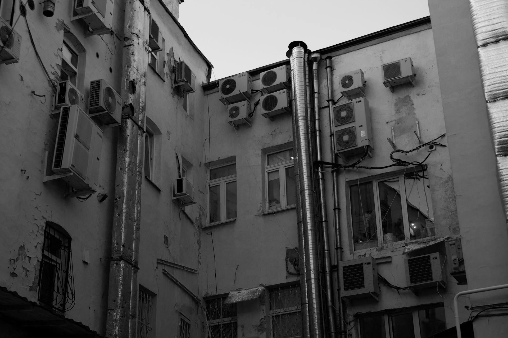 A black and white image of urban building exterior with multiple air conditioning units and pipes.