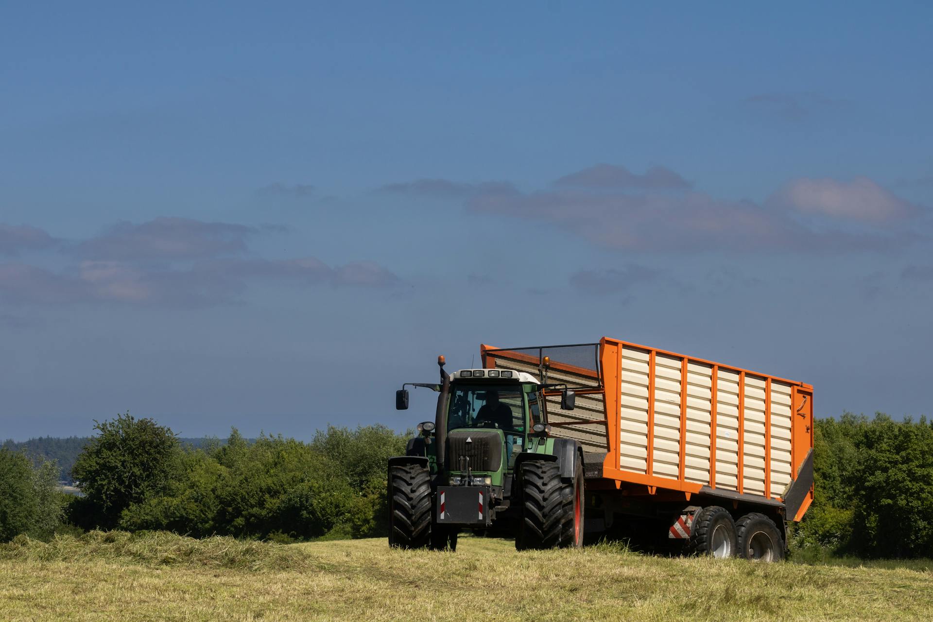 A Fendt tractor with a large trailer working in a lush countryside field under a clear blue sky.