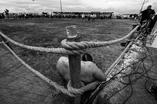 Black and white image of a festival showing a sitting shirtless person behind rope fence.