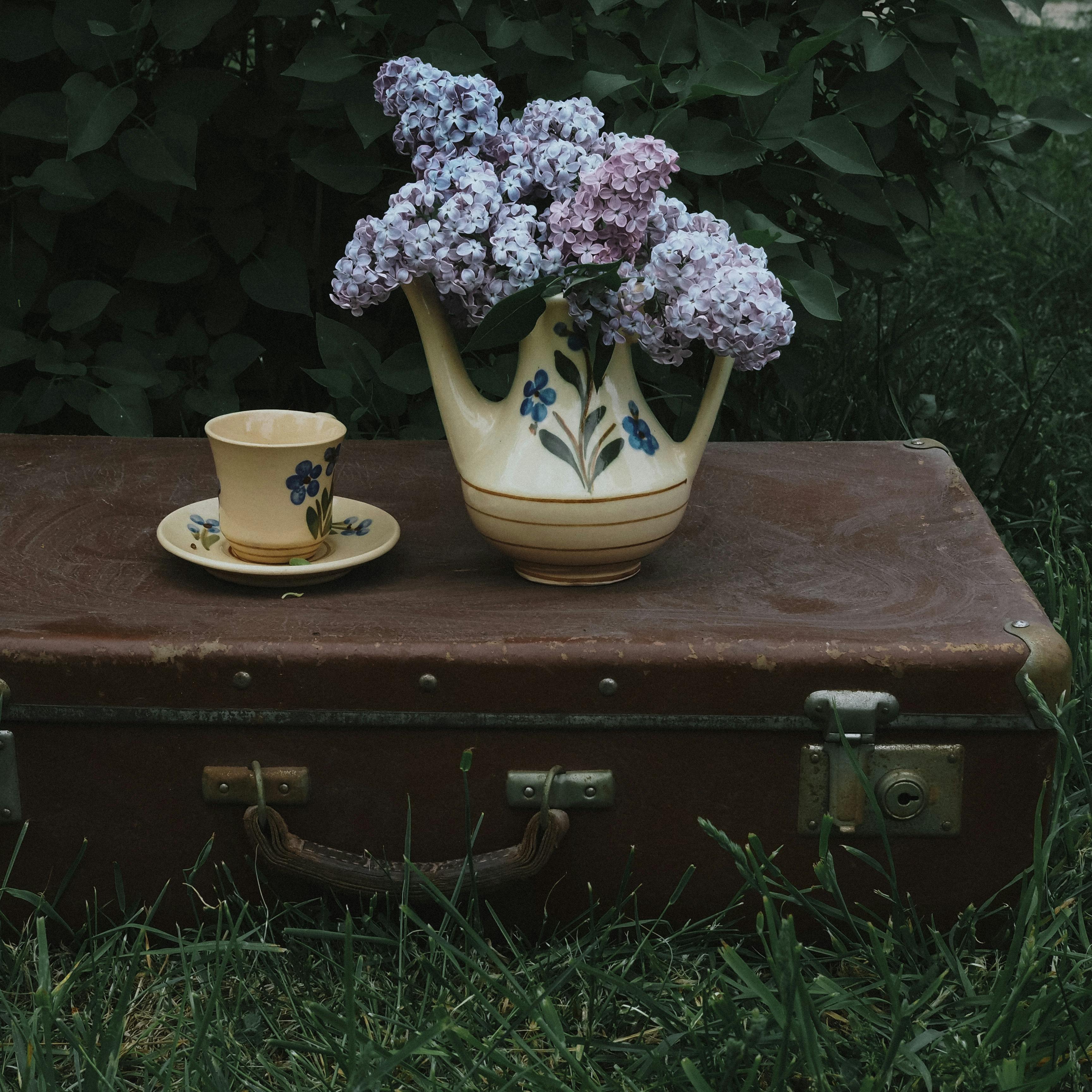 Elegant lilac arrangement in a vintage teapot on a retro suitcase in a garden setting.