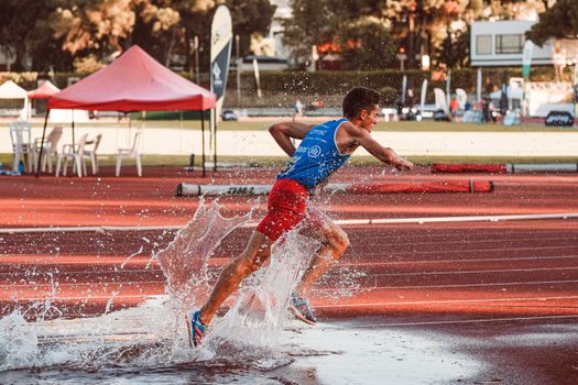 Male athlete running through water splash on track field during steeplechase event.
