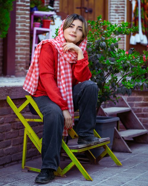 Free Portrait of a young woman in Iran sitting on a chair outdoors, wearing a red outfit with a checkered headscarf. Stock Photo