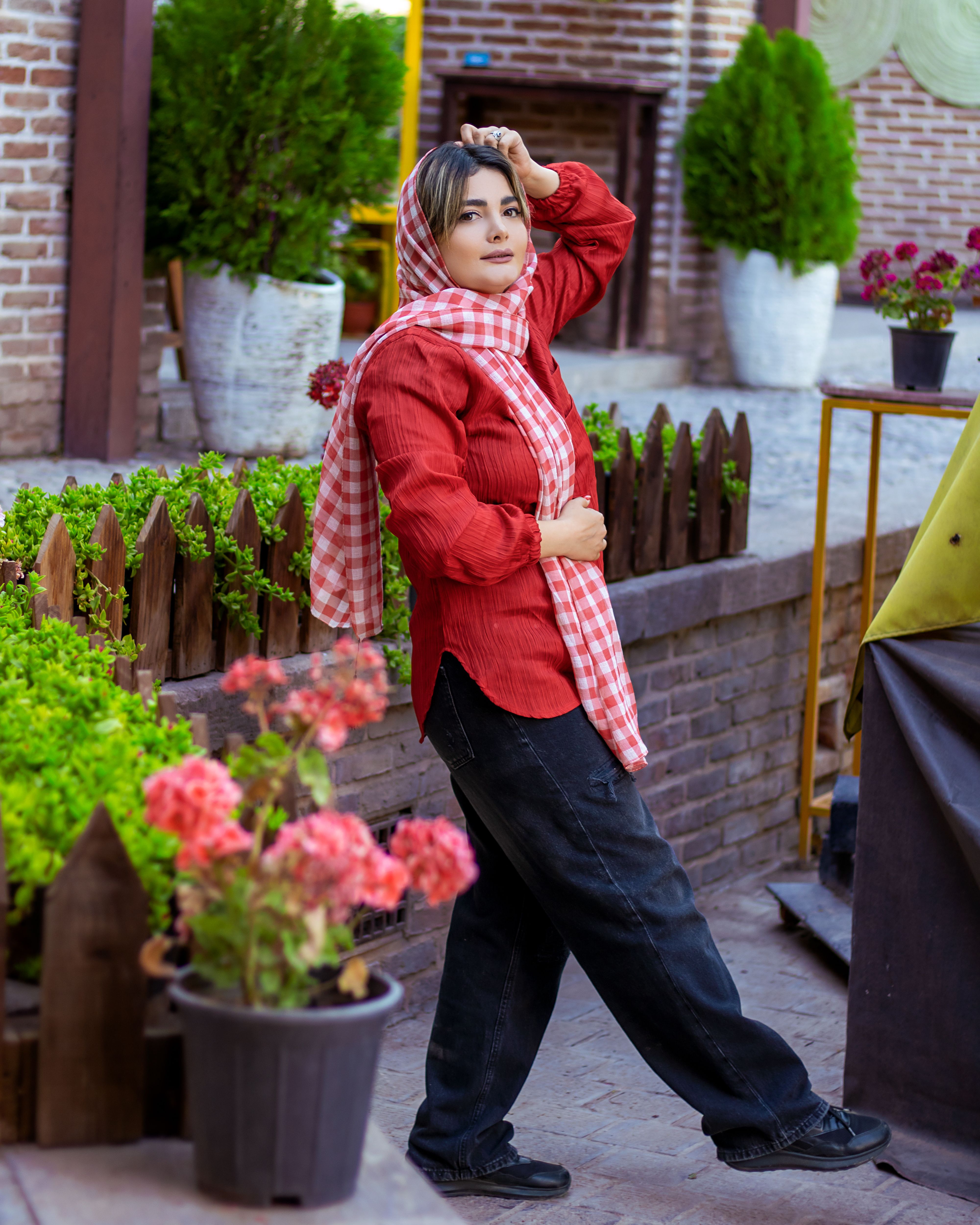 Stylish woman in red shirt and shawl poses confidently in urban garden.