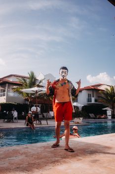 Man in mask and swimming shorts standing poolside on a sunny day, creating a fun and mysterious vibe.