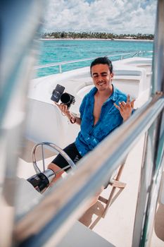 A young man with a camera enjoys a sunny day on a yacht in turquoise waters, capturing moments.