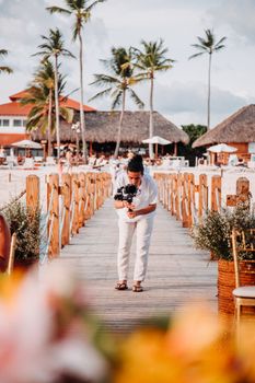 A filmmaker captures scenes on a tropical beach pier, framed by palm trees and thatched huts.