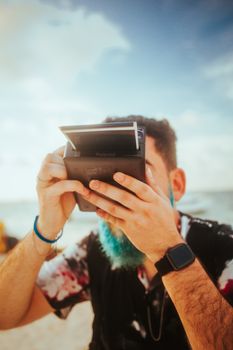 A man with a blue beard uses an instant camera on a sunny beach day.
