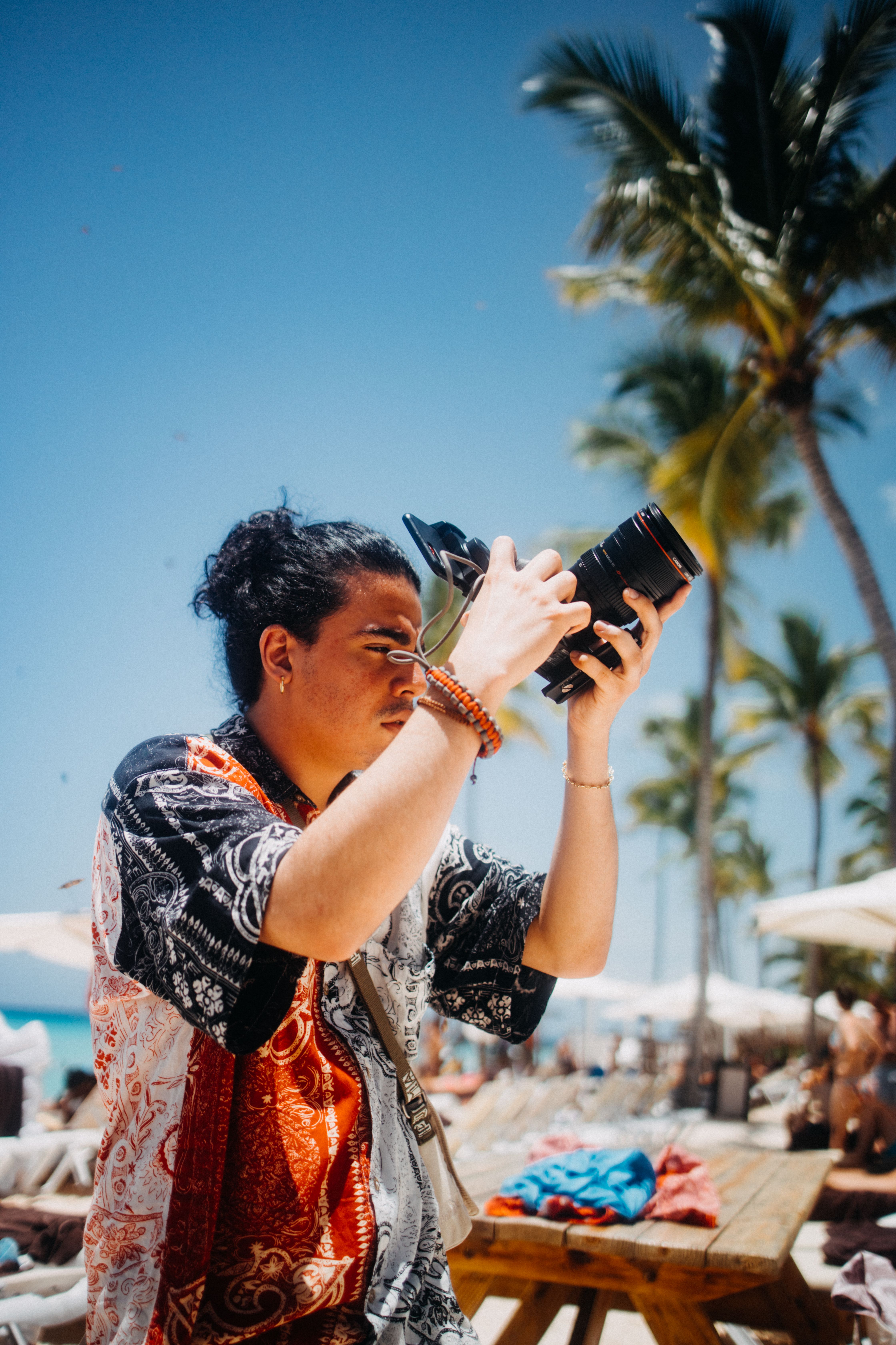 Man with black hair photographing outdoors by the beach under a clear blue sky.