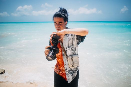 A young photographer with a colorful shirt holds a camera by a tropical ocean beach.