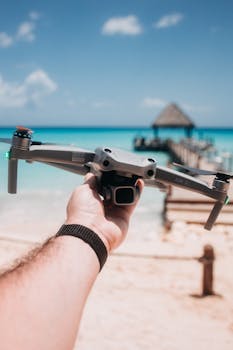 Close-up of a person holding a drone above a sandy beach with turquoise water.