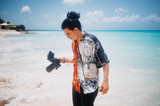 A young adult photographer holding a camera, standing on a sunny tropical beach with turquoise water.