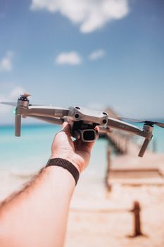 A hand holds a drone by the pier on a sunny beach day, ready for flight.