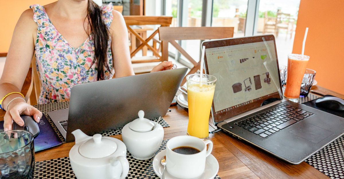 A woman working on laptops at a cafe with drinks and teapots, showcasing a remote work lifestyle.