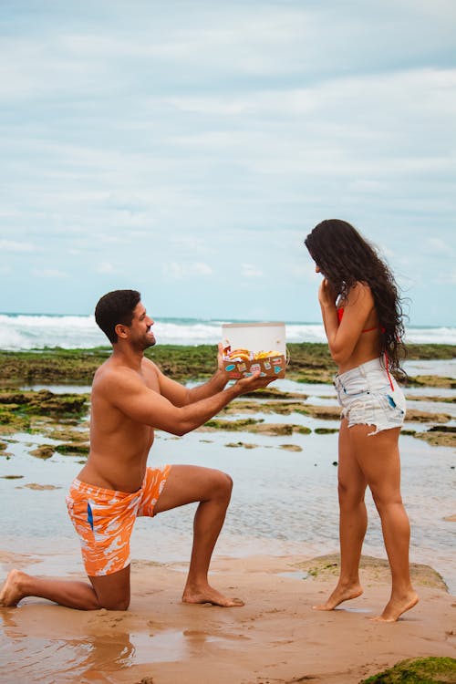 Topless Man in Orange and White Shorts Kneeling in Front of a Woman in Red Bra