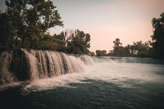 A gentle waterfall cascading under a clear sky surrounded by lush green trees at dusk.