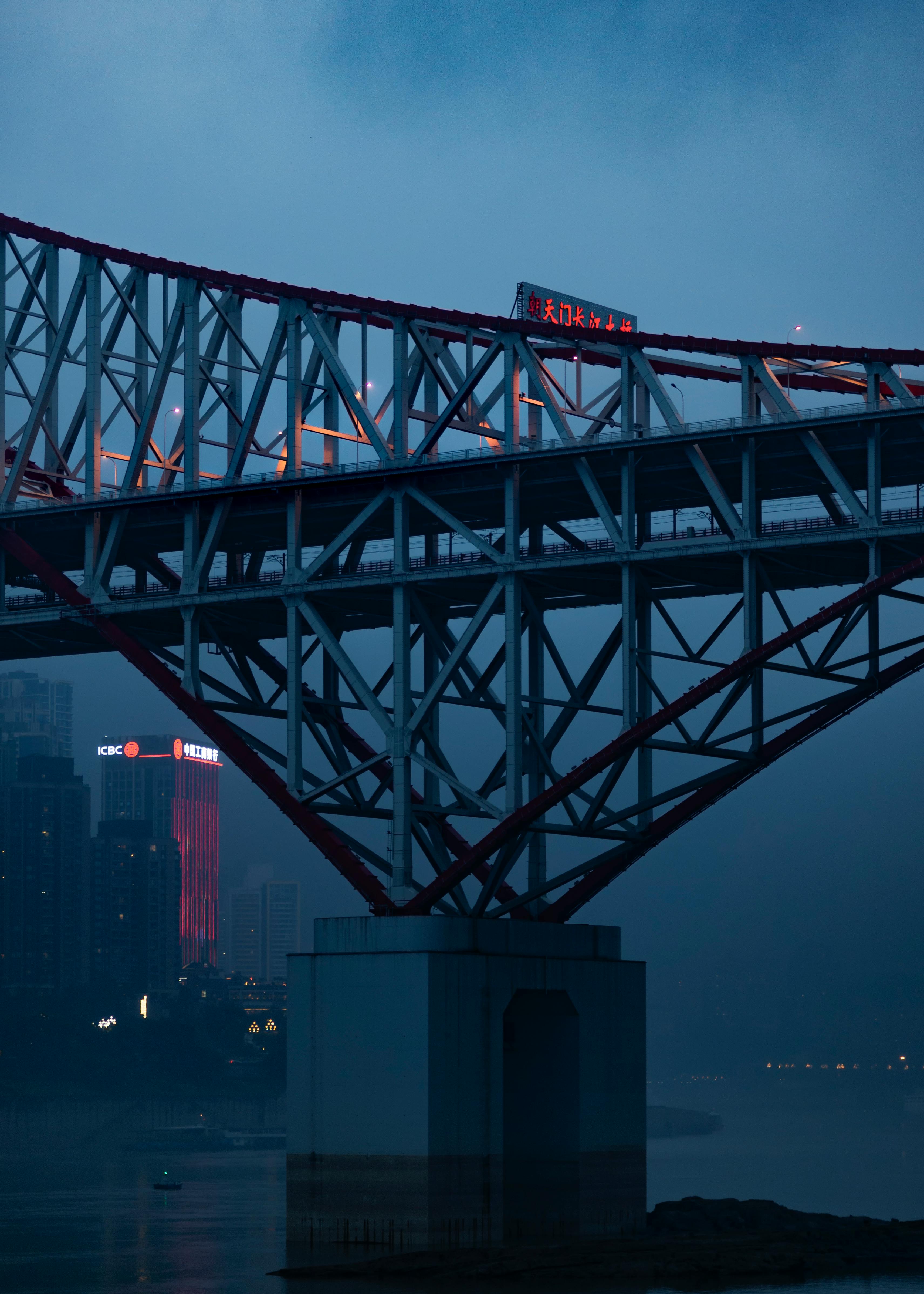 A steel bridge spans a foggy river in an urban setting at dusk with city lights in the background.